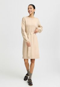 Beiges Strickkleid mit langen Ärmeln und einem Gürtel in der Taille. Es hat einen runden Ausschnitt und eine schlanke Silhouette. Kombiniert mit schwarzen Schuhen und grauen Socken.
