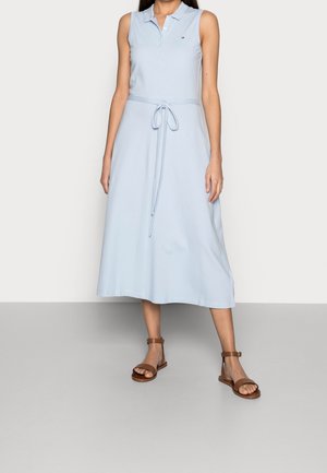 Femme portant une robe midi bleu clair sans manches avec un col et une ceinture nouée, associée à des sandales marron à bride de cheville, debout sur un sol gris.