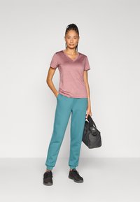 Μπλούζα με λαιμόκοψη V, κοντά μανίκια, σε μπορντό χρώμα με απαλό ύφασμα, συνδυασμένη με τουρκουάζ φόρμες και μαύρα αθλητικά παπούτσια, κρατώντας μια μαύρη τσάντα duffel.