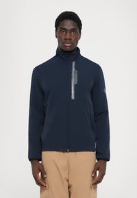 Tamsiai mėlynas zip-up džemperis su aukšta apykakle, turintis atspindinčią juostą ant krūtinės ir siuvinėta logotipu ant rankovės.