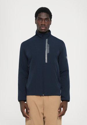 Veste zippée bleu foncé avec un col haut, dotée d'un panneau réfléchissant sur la poitrine et d'un logo brodé sur la manche.