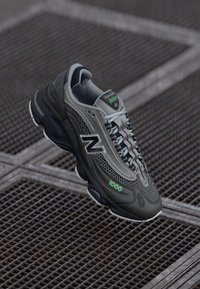 Sivi i crni New Balance 1000 tenisicama s zelenim akcentima, prikazani na metalnoj rešetkastoj podlozi.