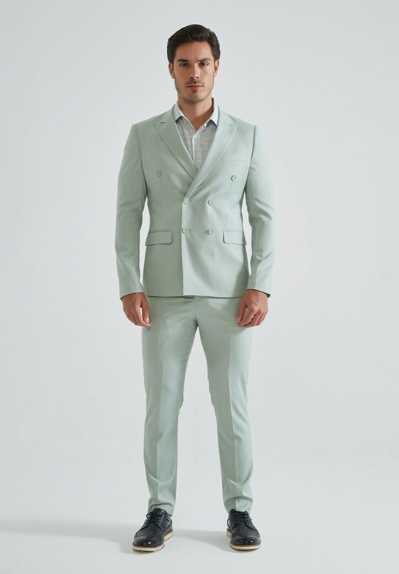 Costume vert clair à double boutonnage et coupe slim, avec des revers en pointe, des poches à rabat et un pantalon assorti, associé à une chemise à motifs et des chaussures noires.