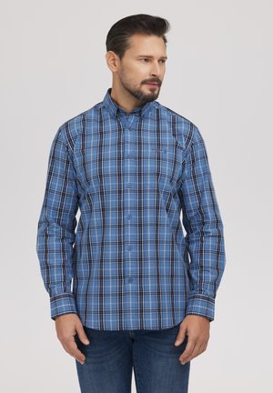 Mężczyzna z krótkimi ciemnymi włosami i brodą, ubrany w niebieską koszulę w kratę na guziki oraz niebieskie dżinsy, stojący na tle jasnego, jednolitego tła.