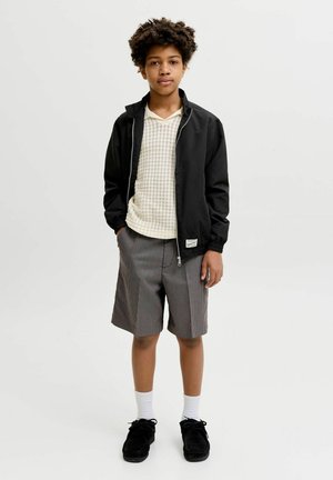 Göndör hajú fiú fekete dzsekiben, fehér kötött ingben, mintás térd fölé érő rövidnadrágban, fehér zokniban és fekete cipőben áll, kezét zsebre dugva.