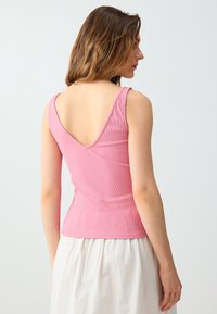 Femme aux cheveux bruns mi-longs portant un haut rose sans manches côtelé avec un dos décolleté et une jupe blanche, de profil.