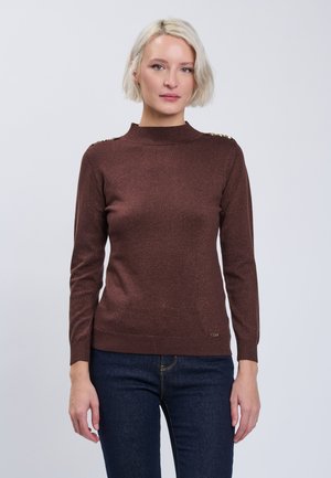 Femme blonde aux cheveux courts portant un pull marron cintré à manches longues et un jean bleu foncé, debout devant un fond clair uni.