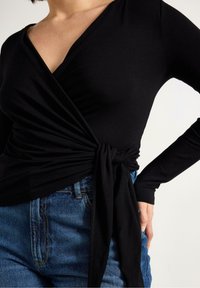 Svart omlottopp med långa ärmar och knytning vid midjan, tillverkad av ett mjukt tyg, kombinerad med högmidjade blå jeans.