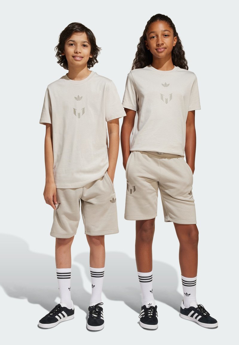 Beige spordikomplekt, mis sisaldab T-särke ja lühikesi pükse, koos delikaatsete logo aktsentidega. Kanga ülesehitus näib olevat pehme ning disain on lihtne ja sportlik.