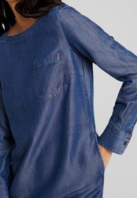 Tummansininen farkk paitapusero, jossa on pyöreä kaula-aukko, pitkät hihat, rintatasku ja napitettu hihansuu; sileä pinta, jossa on kevyt kiilto.