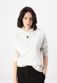 Krémová bavlněná mikina s klokankou a kapucí na šňůrku, s malým černým logem na hrudi. Stylizováno s černými kalhotami.
