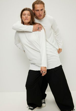 Weiße Langarmoberteile mit geripptem Muster, kombiniert mit lockeren schwarzen Hosen. Beide tragen lässige schwarze Turnschuhe und silberne Ringe.