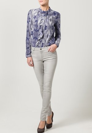 Blusa de manga larga con estampado azul y blanco, con detalle de volantes en el cuello, combinada con pantalones slim-fit en gris claro y tacones oscuros.
