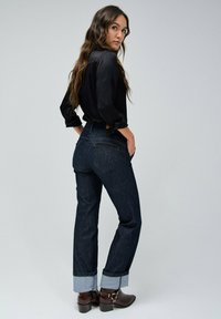 Jeans de mezclilla azul oscuro con puños enrollados, cintura alta y detalles de costura sutiles, combinados con una camiseta de manga larga negra y botas marrones.