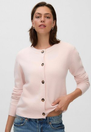 Femme aux cheveux bruns courts porte un cardigan rose clair boutonné et un jean bleu, tenant le bord inférieur du cardigan d’une main.