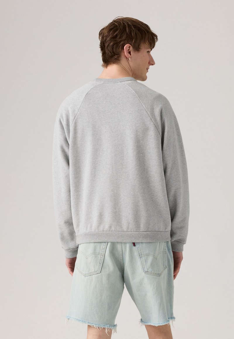 Sudadera gris con cuello redondo, mangas raglán, puños y cinturilla acanalados. Lleva puesta con pantalones cortos de mezclilla azul claro con bordes deshilachados.