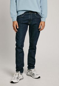 Jeans de mezclilla azul oscuro con corte recto, que presentan un clásico diseño de cinco bolsillos y sutilezas en la costura, combinados con zapatillas blancas.