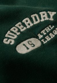 Tissu vert avec texte blanc brodé disant "SUPERDRY 1ST ATHL LEAG" en style arrondi et lettres majuscules.