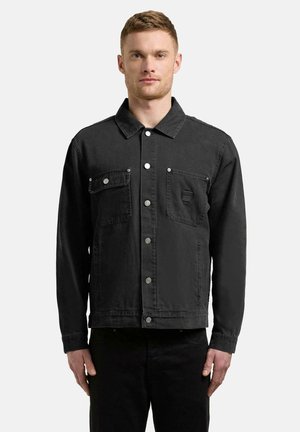 Unge mann iført en svart denimjakke med knapper og to brystlommer, stående foran en ensfarget lys grå bakgrunn.