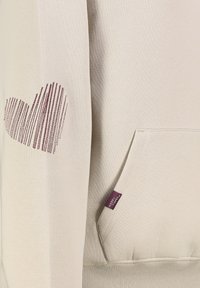 Beige sweatshirt met een stevige textuur, featuring een bordeauxrood hartontwerp op de linkermouw en een zak aan de voorkant. Label zichtbaar aan de zijkant.