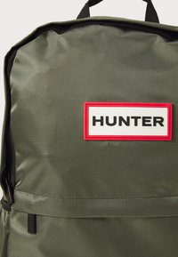 Mochila de tela verde oliva con cremallera y asa negras, que presenta un parche blanco con borde rojo y el texto "HUNTER" en negrita negra.