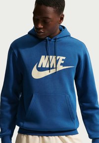 Junger Mann trägt blauen Nike-Hoodie mit Fronttasche und großem weißem Logo, blickt nach unten, vor einfachem hellem Hintergrund.