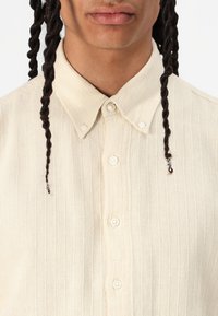Lichtbeige knoopshirt met een textuur van ribstof. Heeft een klassieke kraag en parelachtige knopen aan de voorkant.