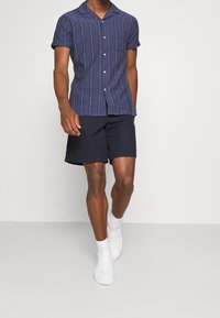 Camicia a maniche corte navy con bottoni, strisce verticali bianche e taschino sul petto, abbinata a pantaloni corti navy e sneaker bianche.