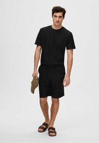 Juodas medvilninis marškinėliai ir šortai, laisvas kirpimas, su lygiu paviršiumi. Taip pat yra rudi sandalai ir smėlio spalvos kepurė.