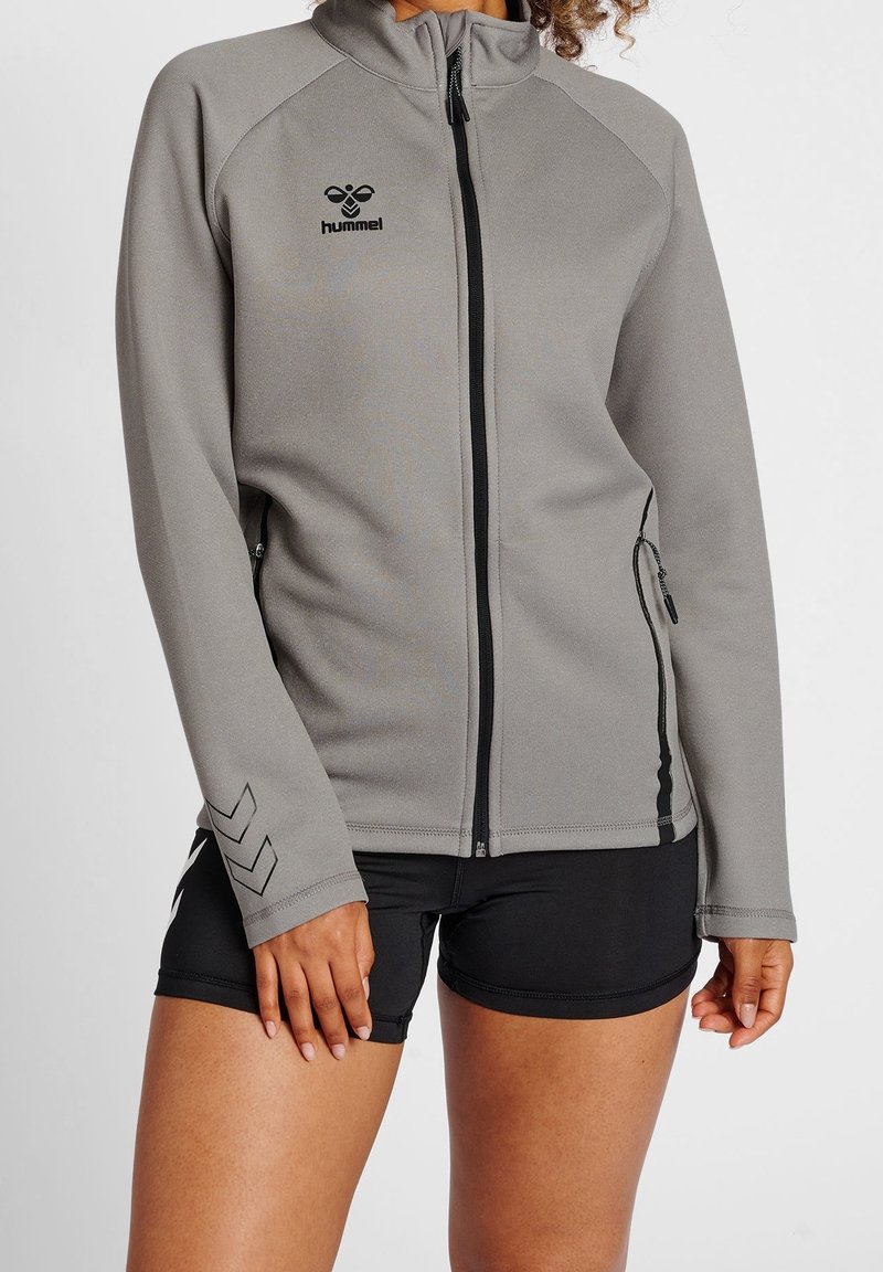 Grå zip-up sportjacka med hög krage, tillverkad av slät tyg. Har svarta detaljer, sidofickor och logotyp på bröstet.