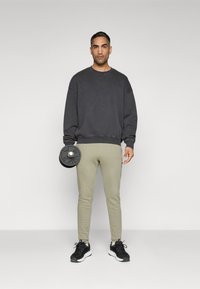 Sweatshirt oversized cinza escuro, calças justas verde-oliva claro, segurando uma placa de peso preta, com tênis pretos com solas brancas.