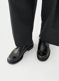 Czarne brogsy ze skóry z ozdobnymi perforacjami i brązowym przeszyciem, w zestawieniu z czarnymi spodniami, prezentujące klasyczny design i wypolerowaną fakturę.