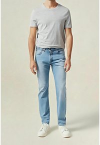 Homme portant un t-shirt gris clair à manches courtes, un jean bleu clair et des baskets blanches, debout devant un fond uni.