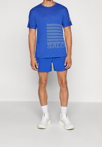 Blå idrottströja med horisontella linjer och texten "SAL"; matchas med blå shorts. Vita sneakers med gröna detaljer.