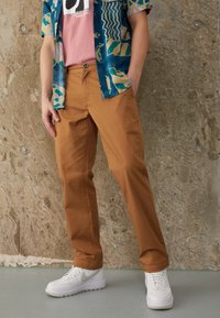 Persoon in een bruine broek, witte sneakers, een roze T-shirt met opdruk en een ongeknopte blauwe geblokte kortemouwen overhemd tegen een gestructureerde muur.