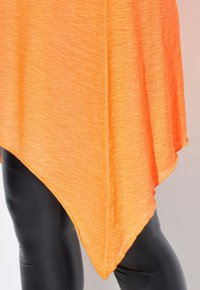Túnica asimétrica naranja con tejido texturizado, que presenta un dobladillo triangular y una caída ligera, combinada con leggings negros.
