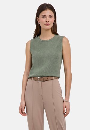 Jeune femme portant un haut en maille vert sans manches, un pantalon beige taille haute avec ceinture, des boucles d'oreilles anneaux et un bracelet à son poignet droit.