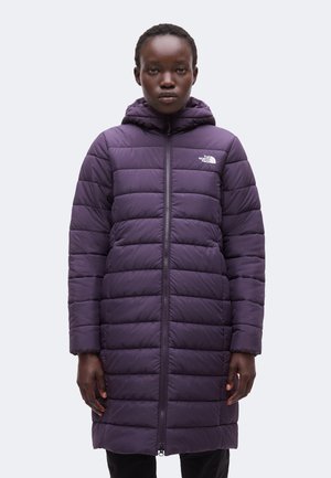 Persona con una chaqueta larga acolchada de color púrpura oscuro con capucha y cremallera frontal, de pie frente a un fondo claro y liso.