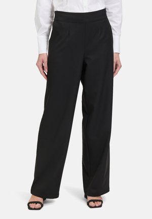 Schwarze, weit geschnittene Hose aus glattem Stoff mit flachem Bund und zwei vorderen Falten, kombiniert mit einem weißen Hemd mit Knöpfen und schwarzen Sandalen.