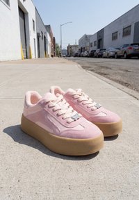 Paire de baskets à plateforme roses avec des semelles épaisses couleur beige, posées sur un trottoir en béton dans un cadre urbain avec des voitures garées.