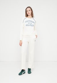 Witte sweatshirt met marineblauwe "Converse All Star" print, gecombineerd met bijpassende lichte broek. Model draagt groene sneakers. Eenvoudig ontwerp, relaxte pasvorm.