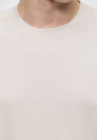 Gros plan sur le cou et le haut de la poitrine d’une personne portant un t-shirt en coton beige clair à col rond.