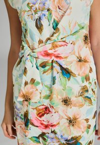 Robe florale avec un fond clair, ornée de grandes fleurs roses, bleues et jaunes. Elle est dotée d'une taille nouée et d'un tissu fluide et texturé.