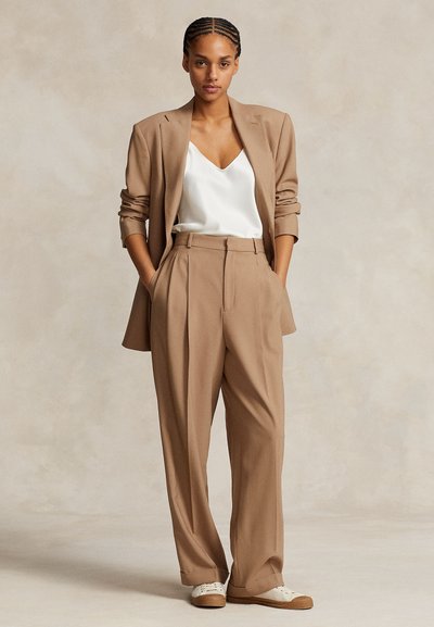 Femme debout les mains dans les poches portant un blazer beige et un pantalon large assorti, un caraco en satin blanc et des baskets blanches.