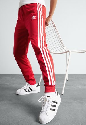 Osoba ubrana w czerwone spodnie dresowe Adidas z białymi paskami i białe buty sportowe Adidas trzyma białe metalowe krzesło na szarej podłodze.