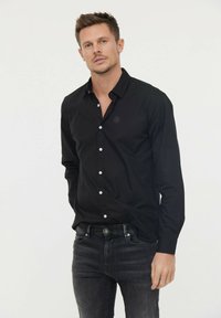 Chemise noire à manches longues avec un col boutonné et des boutons blancs, dotée d'un petit logo sur la poitrine et d'un tissu lisse et léger.