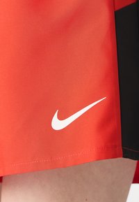 Červené šortky Nike s čiernymi akcentmi, jemná tkanina, rovný strih a výrazné biele logo Nike swoosh blízko dolného okraja.