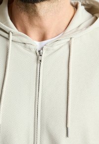 Světlá béžová mikina na zip s texturovanou tkaninou, vybavená stříbrným zipem a šňůrkami pro stažení. Detailní záběr na oblast krku.