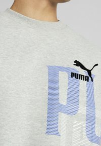 Sudadera gris claro con gran texto "PUMA" en azul y blanco con puntos, logo negro y cuello redondo acanalado. Tejido suave de mezcla de algodón.