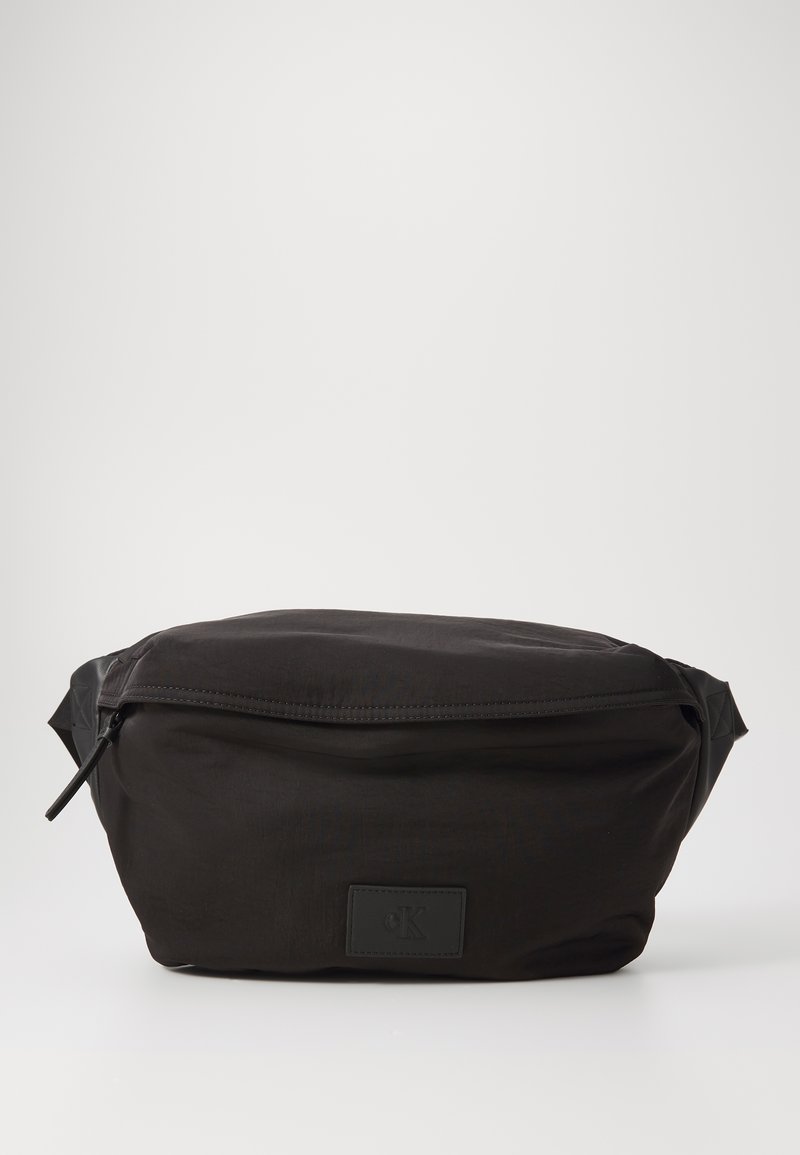 Marsupio in nylon nero con zip nella parte superiore, dotato di un'etichetta logo rettangolare. Il marsupio ha una superficie liscia e un design minimalista.
